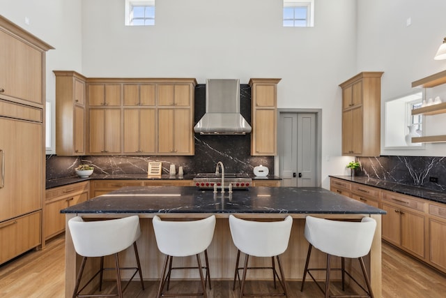 kitchen featuring an island with sink, wall chimney exhaust hood, paneled built in refrigerator, a high ceiling, and light wood-style floors