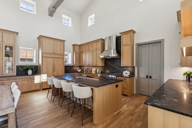 kitchen featuring paneled refrigerator, a sink, light wood-style floors, wall chimney range hood, and an island with sink