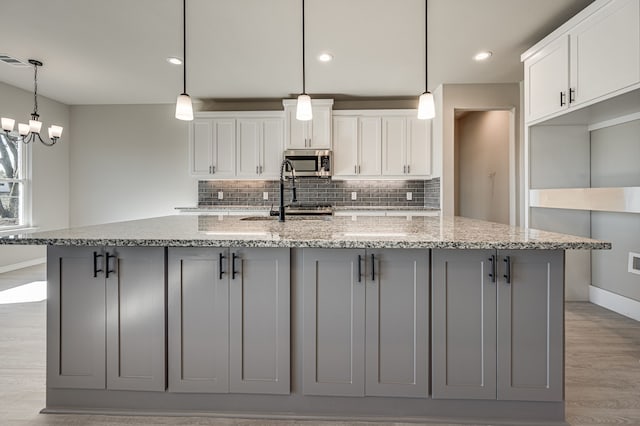 kitchen featuring white cabinets, light stone countertops, stainless steel microwave, decorative light fixtures, and an island with sink