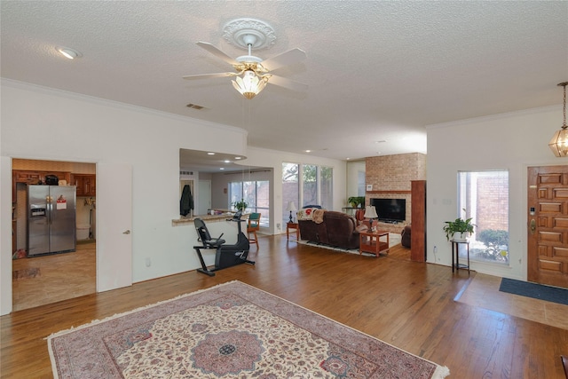 living room featuring a textured ceiling, wood finished floors, visible vents, a brick fireplace, and crown molding