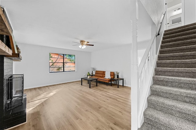 living area featuring light wood-style flooring, stairway, a ceiling fan, a brick fireplace, and baseboards