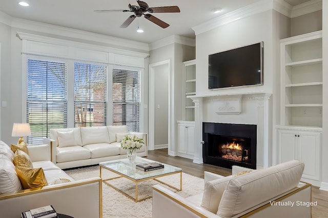 living room featuring built in shelves, crown molding, a warm lit fireplace, wood finished floors, and a ceiling fan