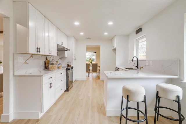 kitchen with a peninsula, under cabinet range hood, stainless steel range with electric stovetop, white cabinetry, and a sink