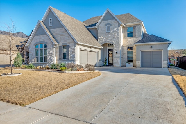 french country inspired facade with a garage, stone siding, a shingled roof, and driveway