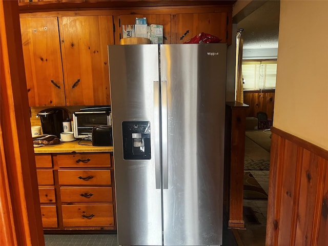 kitchen with wooden walls, wainscoting, brown cabinetry, and stainless steel refrigerator with ice dispenser