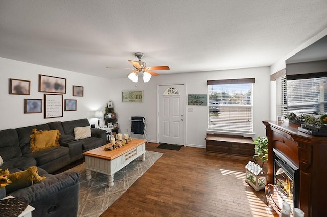 living room with a warm lit fireplace, ceiling fan, dark wood-type flooring, and baseboards