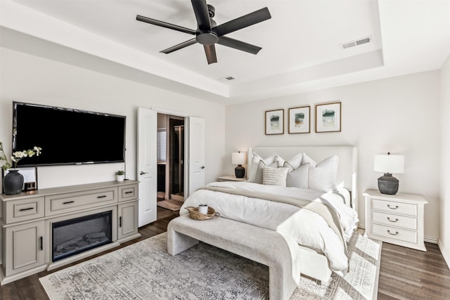 bedroom with dark wood-style floors, a tray ceiling, a glass covered fireplace, and visible vents