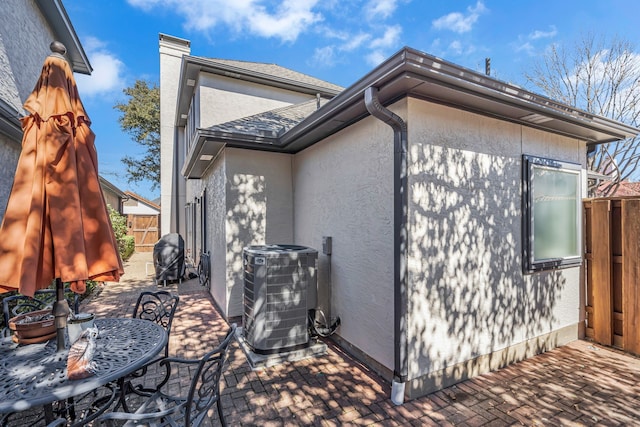 view of side of property with a patio, central AC, a shingled roof, fence, and stucco siding