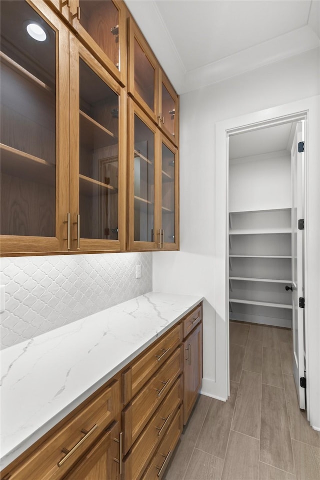 kitchen with tasteful backsplash, glass insert cabinets, brown cabinetry, ornamental molding, and light stone countertops