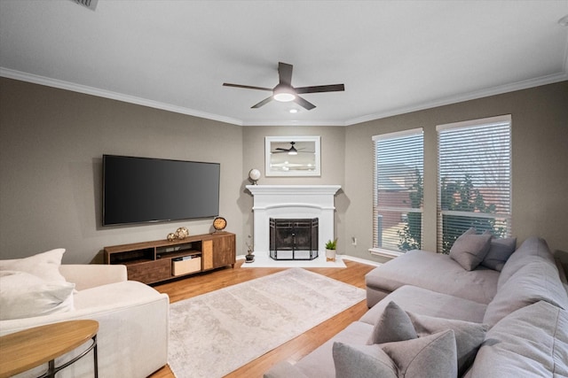 living room featuring ceiling fan, light wood-style flooring, a fireplace with flush hearth, baseboards, and ornamental molding