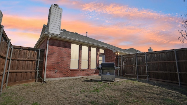 back of property at dusk with brick siding, a chimney, a fenced backyard, and a lawn