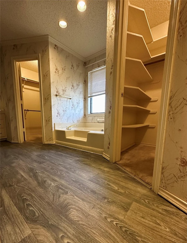 interior space with a walk in closet, a garden tub, ornamental molding, a textured ceiling, and wallpapered walls