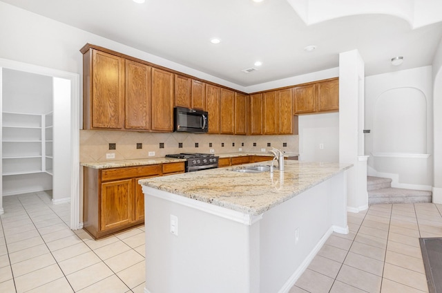 kitchen with an island with sink, light stone counters, brown cabinets, stainless steel range with gas cooktop, and a sink