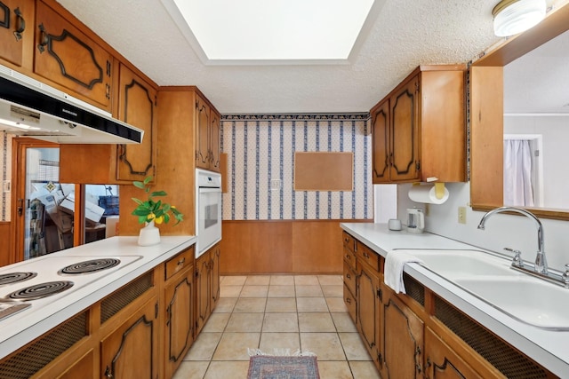 kitchen with wallpapered walls, brown cabinetry, a textured ceiling, under cabinet range hood, and a sink