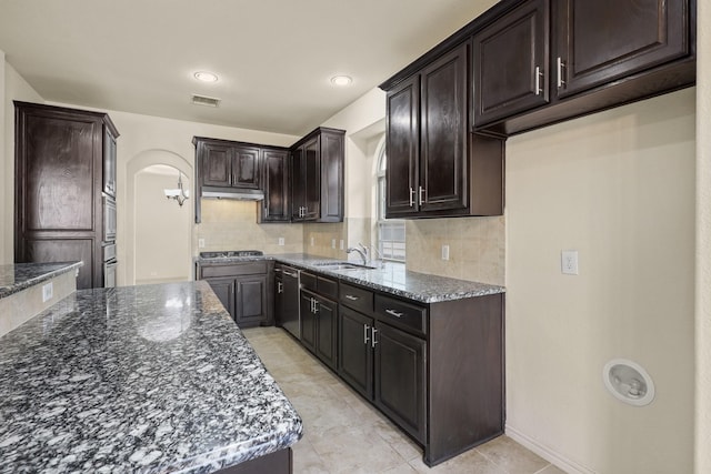 kitchen with arched walkways, under cabinet range hood, stainless steel gas cooktop, a sink, and dark stone counters