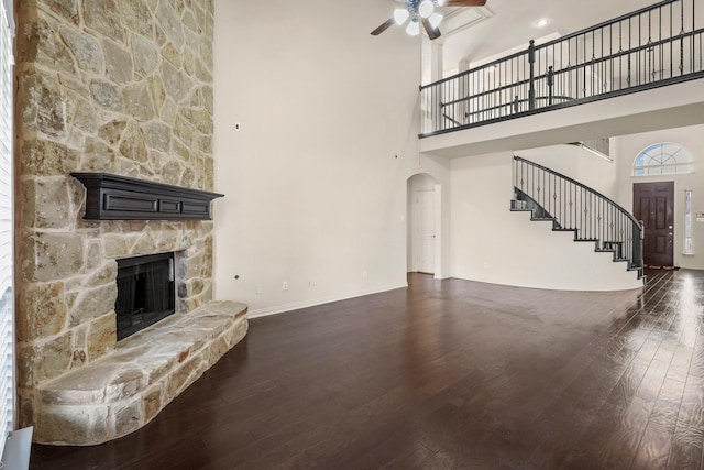 unfurnished living room featuring arched walkways, a stone fireplace, dark wood-type flooring, a ceiling fan, and stairs