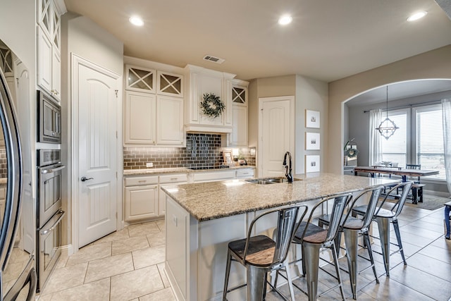 kitchen featuring arched walkways, a kitchen island with sink, a sink, white cabinetry, and glass insert cabinets