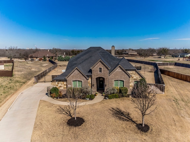 french country inspired facade with driveway, a shingled roof, a chimney, fence, and brick siding