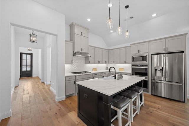 kitchen featuring a kitchen island with sink, under cabinet range hood, appliances with stainless steel finishes, light stone countertops, and a kitchen bar