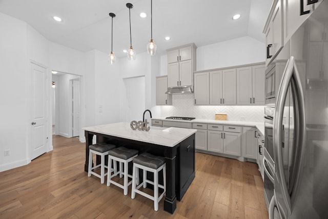 kitchen featuring under cabinet range hood, stainless steel appliances, a sink, hanging light fixtures, and a center island with sink