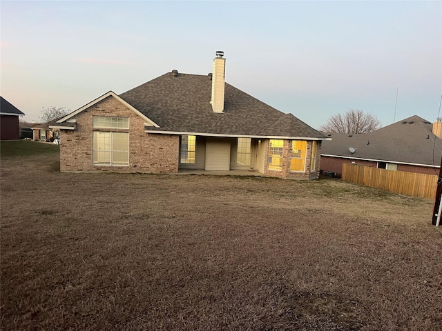 back of house at dusk with brick siding, a chimney, a shingled roof, a lawn, and fence