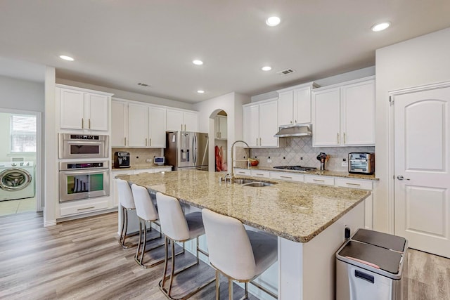 kitchen featuring arched walkways, appliances with stainless steel finishes, washer / clothes dryer, under cabinet range hood, and a sink