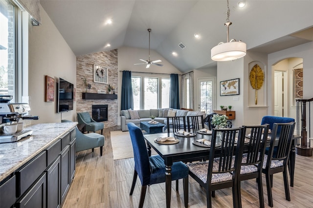 dining area with light wood finished floors, visible vents, arched walkways, a stone fireplace, and high vaulted ceiling