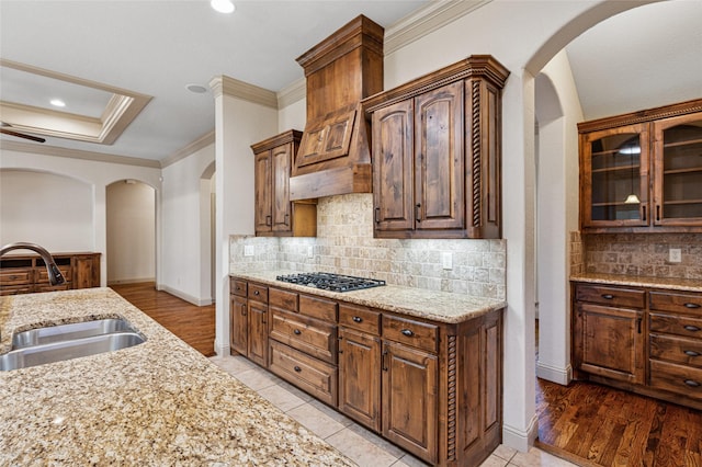 kitchen with glass insert cabinets, light stone counters, ornamental molding, gas stovetop, and a sink