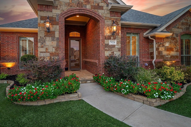 exterior entry at dusk with stone siding, brick siding, and roof with shingles