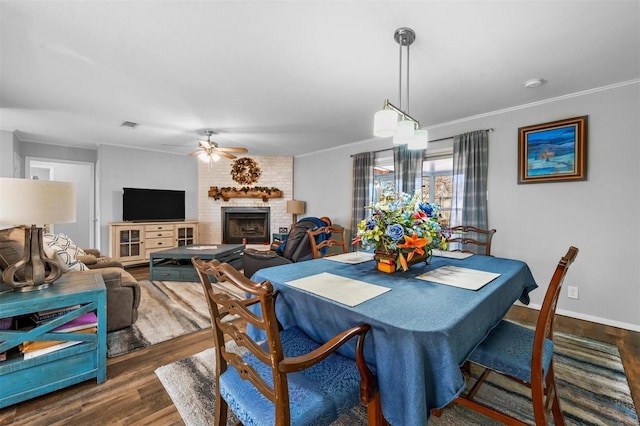 dining room with crown molding, dark wood finished floors, visible vents, a ceiling fan, and a brick fireplace