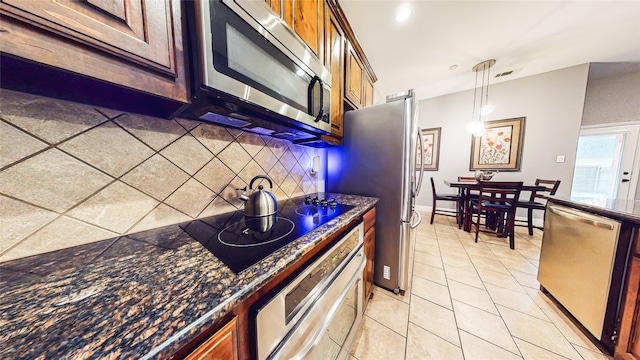 kitchen with light tile patterned floors, dark stone counters, appliances with stainless steel finishes, hanging light fixtures, and backsplash