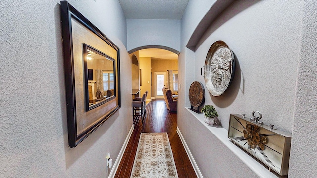 hall featuring baseboards, arched walkways, dark wood-style flooring, and a textured wall