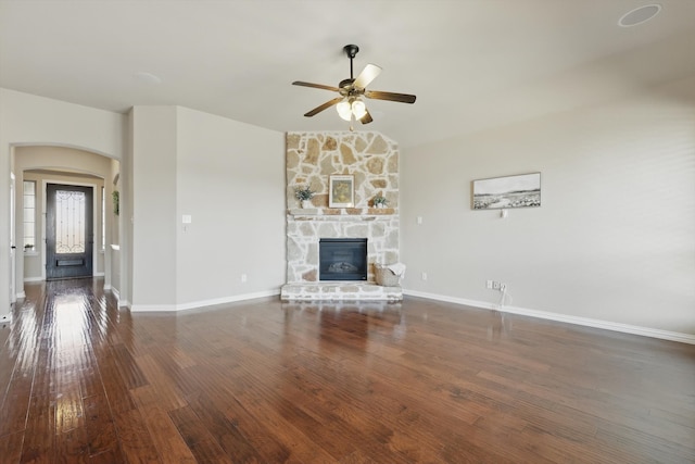 unfurnished living room with arched walkways, a stone fireplace, hardwood / wood-style flooring, a ceiling fan, and baseboards