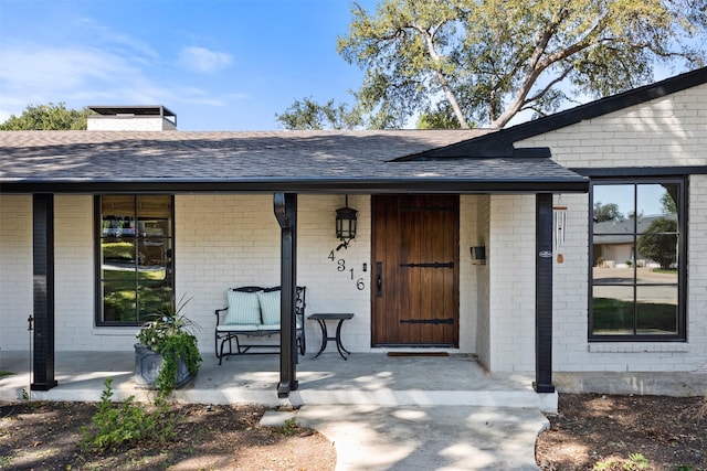 doorway to property featuring covered porch, a chimney, brick siding, and roof with shingles