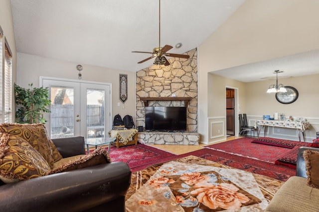 living room with french doors, wainscoting, vaulted ceiling, a stone fireplace, and ceiling fan with notable chandelier