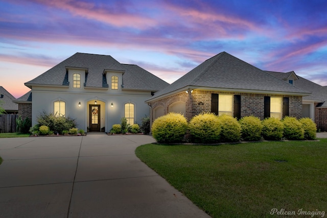 french country style house with driveway, a garage, a shingled roof, a yard, and brick siding