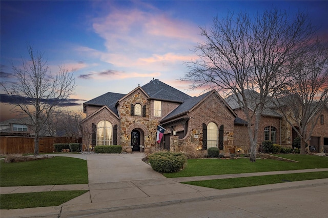 french provincial home with concrete driveway, stone siding, fence, a front lawn, and brick siding