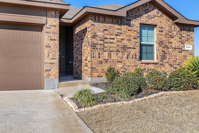 entrance to property featuring a garage, brick siding, and a shingled roof