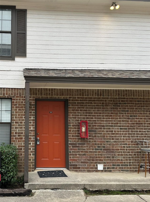 view of exterior entry featuring a shingled roof and brick siding