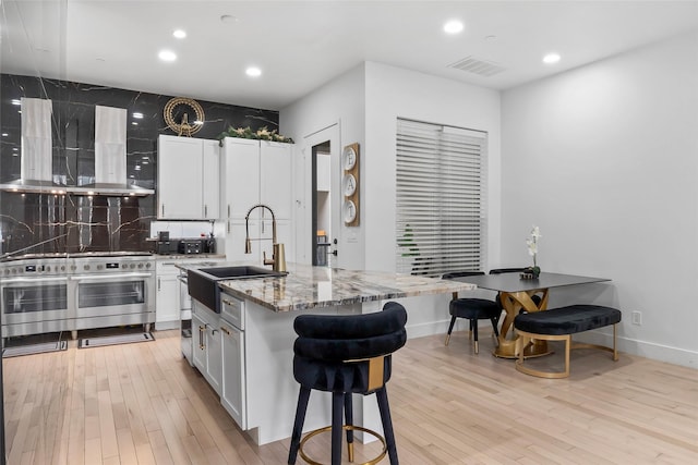 kitchen featuring white cabinets, range with two ovens, an island with sink, a sink, and exhaust hood