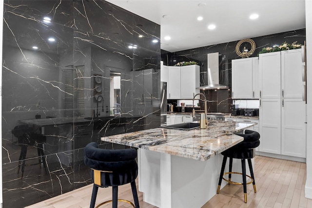 kitchen featuring light stone counters, a breakfast bar, white cabinets, wall chimney range hood, and an island with sink