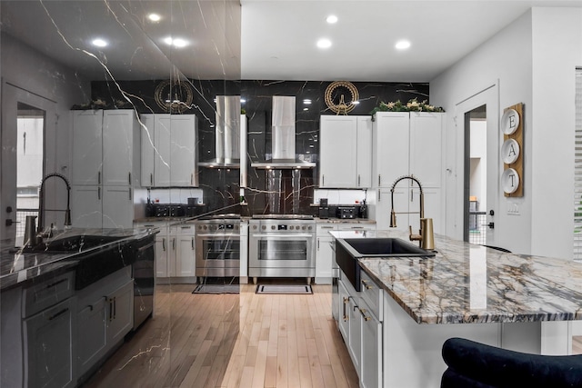 kitchen featuring stone countertops, a center island with sink, white cabinets, high end stainless steel range oven, and wall chimney range hood