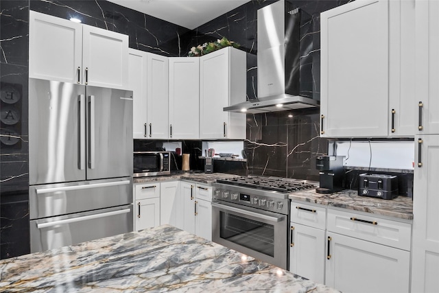 kitchen featuring stainless steel appliances, wall chimney range hood, white cabinetry, and light stone countertops