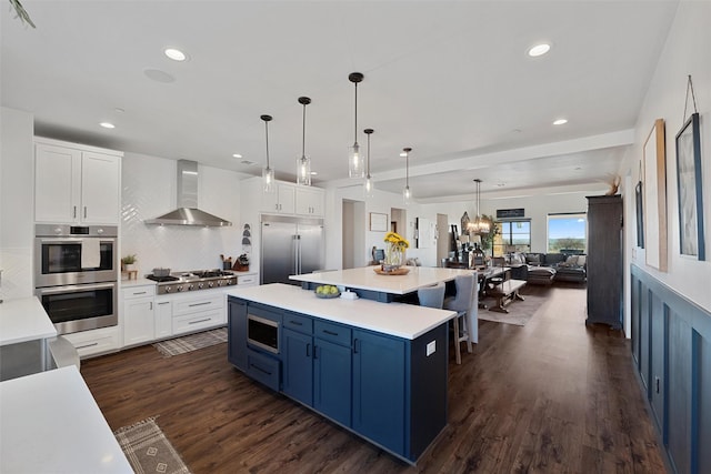 kitchen with built in appliances, white cabinetry, light countertops, blue cabinetry, and wall chimney exhaust hood