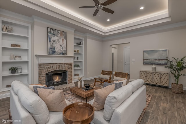 living area featuring built in shelves, a tray ceiling, dark wood-style flooring, a brick fireplace, and baseboards