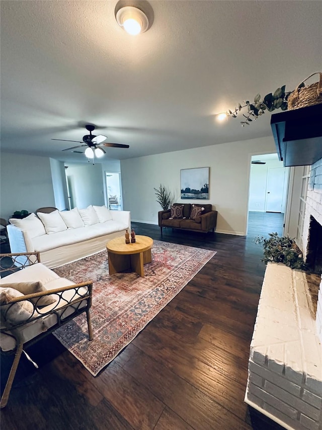 living room with a textured ceiling, ceiling fan, dark wood-style flooring, baseboards, and a brick fireplace