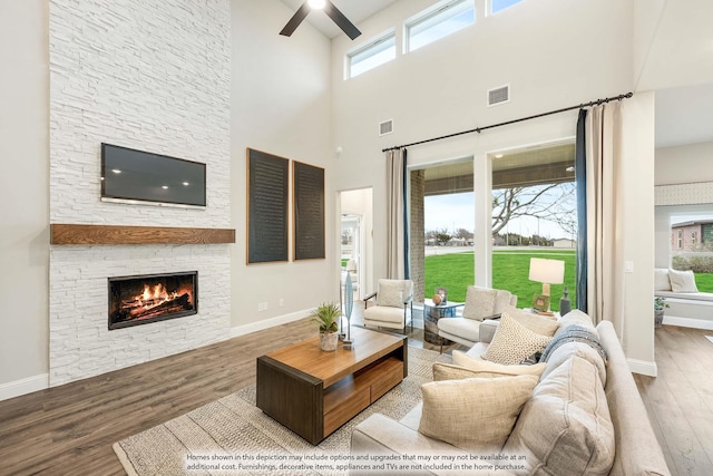 living room with baseboards, visible vents, a ceiling fan, wood finished floors, and a stone fireplace