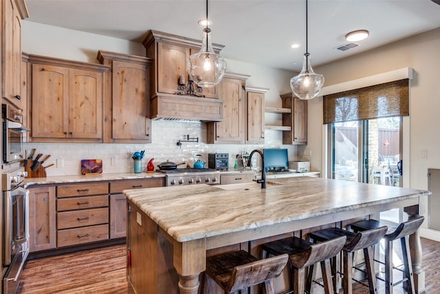 kitchen featuring a center island with sink, range, light stone counters, hanging light fixtures, and a sink