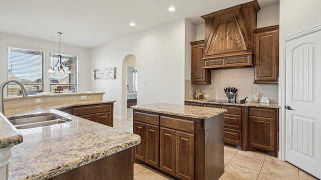 kitchen with a center island with sink, hanging light fixtures, decorative backsplash, a sink, and premium range hood