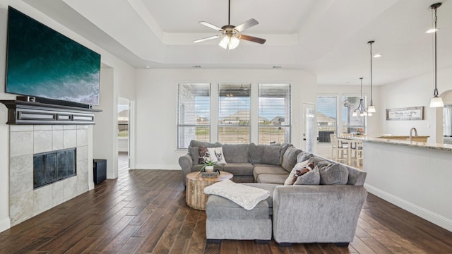 living area featuring ceiling fan with notable chandelier, a fireplace, baseboards, dark wood-style floors, and a raised ceiling